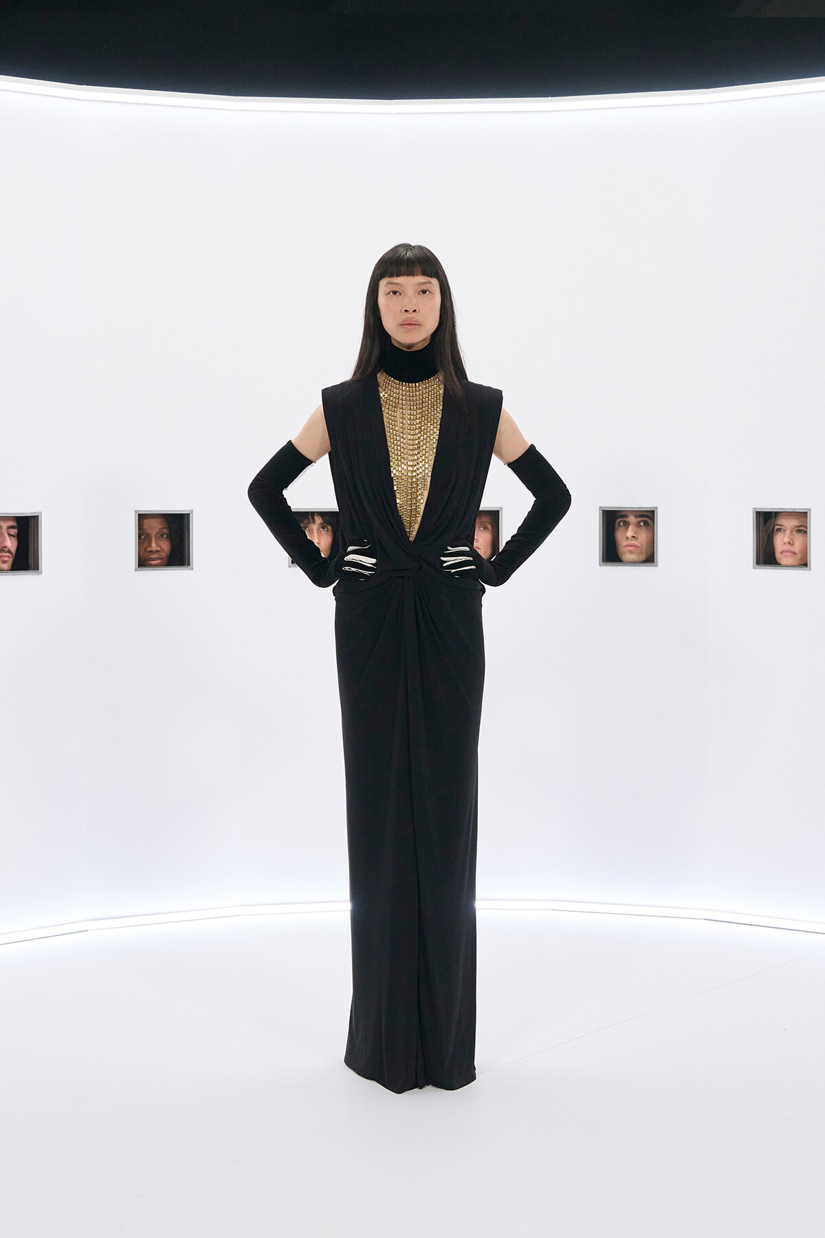 Model in a black, floor-length gown with a gold beaded bib, standing with hands on hips in a bright white studio with small square window portraits along the wall.