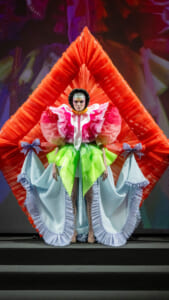 Model on a runway wearing a neon green, sculptural skirt with a pink-and-white ruffled bodice, a pale blue cape with bows, against a large red geometric backdrop.