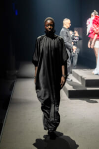 Model walks a black satin cape dress on a dimly lit runway, head covered, spotlight on stage.