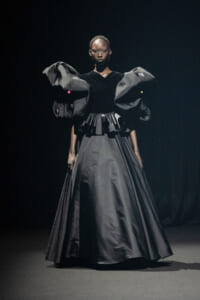 Female model in a dramatic, floor-length black gown with oversized sculptural sleeves on a dark runway setting.