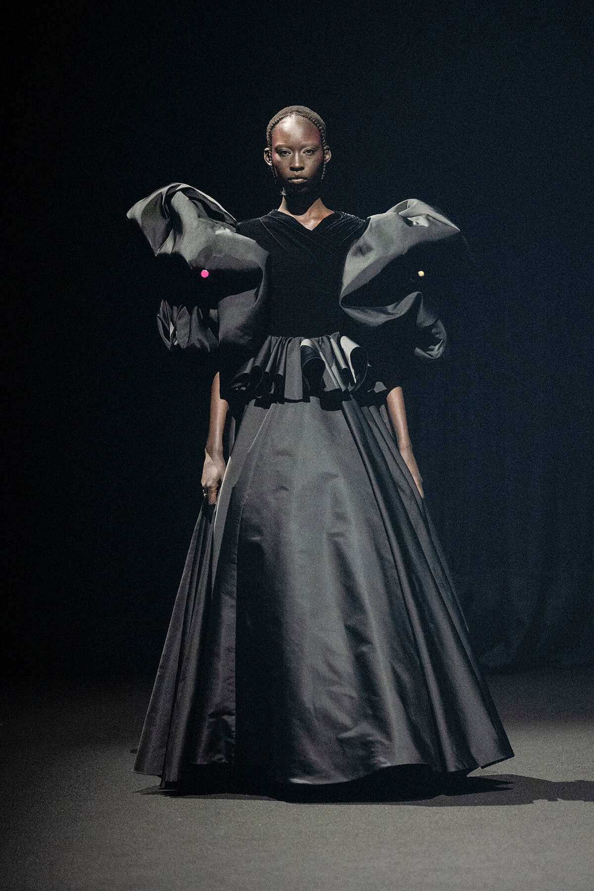 Female model in a dramatic, floor-length black gown with oversized sculptural sleeves on a dark runway setting.