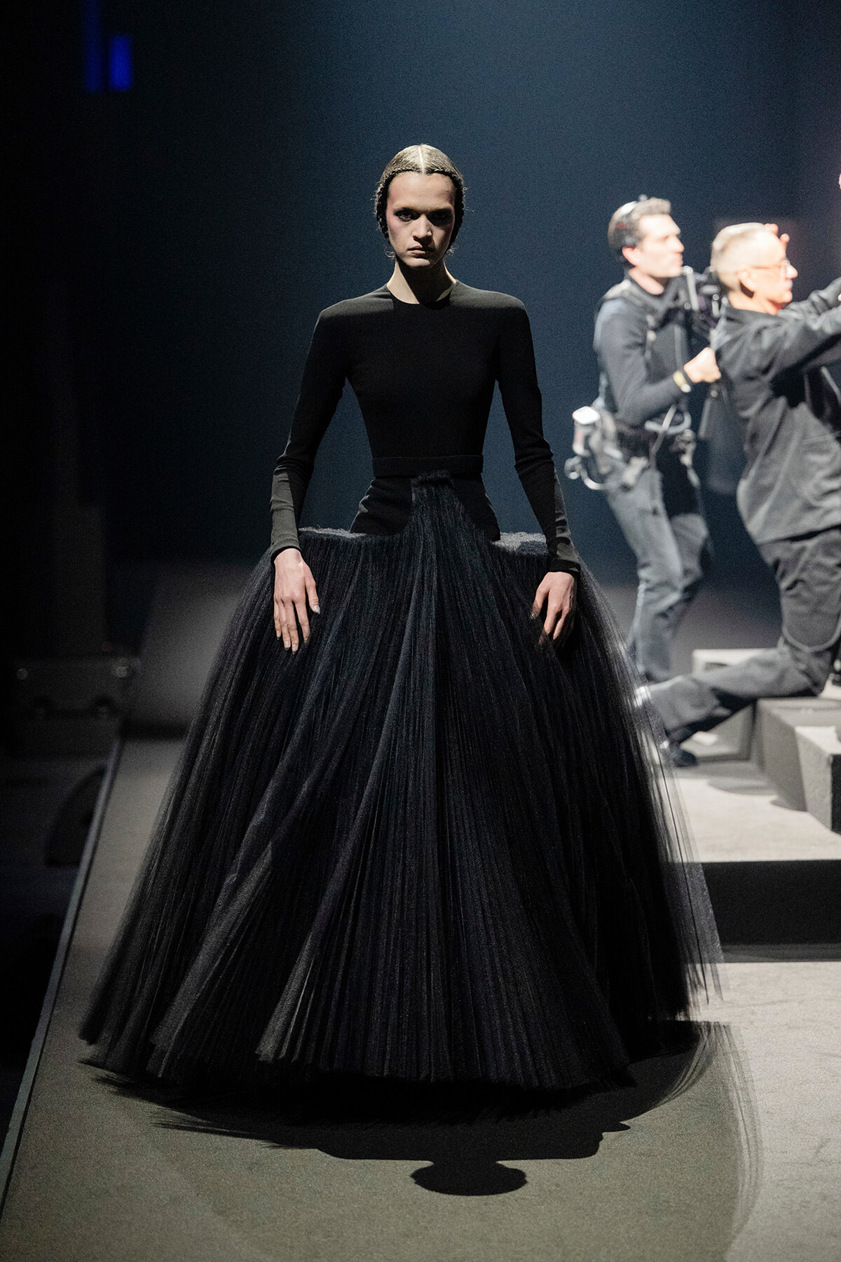 Model on a runway wearing an all-black outfit: a fitted long-sleeve top and a voluminous black tulle skirt with a dramatic silhouette.
