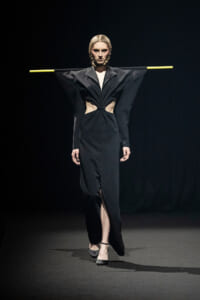 Model walking a fashion show in a black, avant-garde gown with wing-like sleeves, cutouts at the waist, and a front slit; hair styled in braids and arms stretched out with yellow bars across the sleeves.