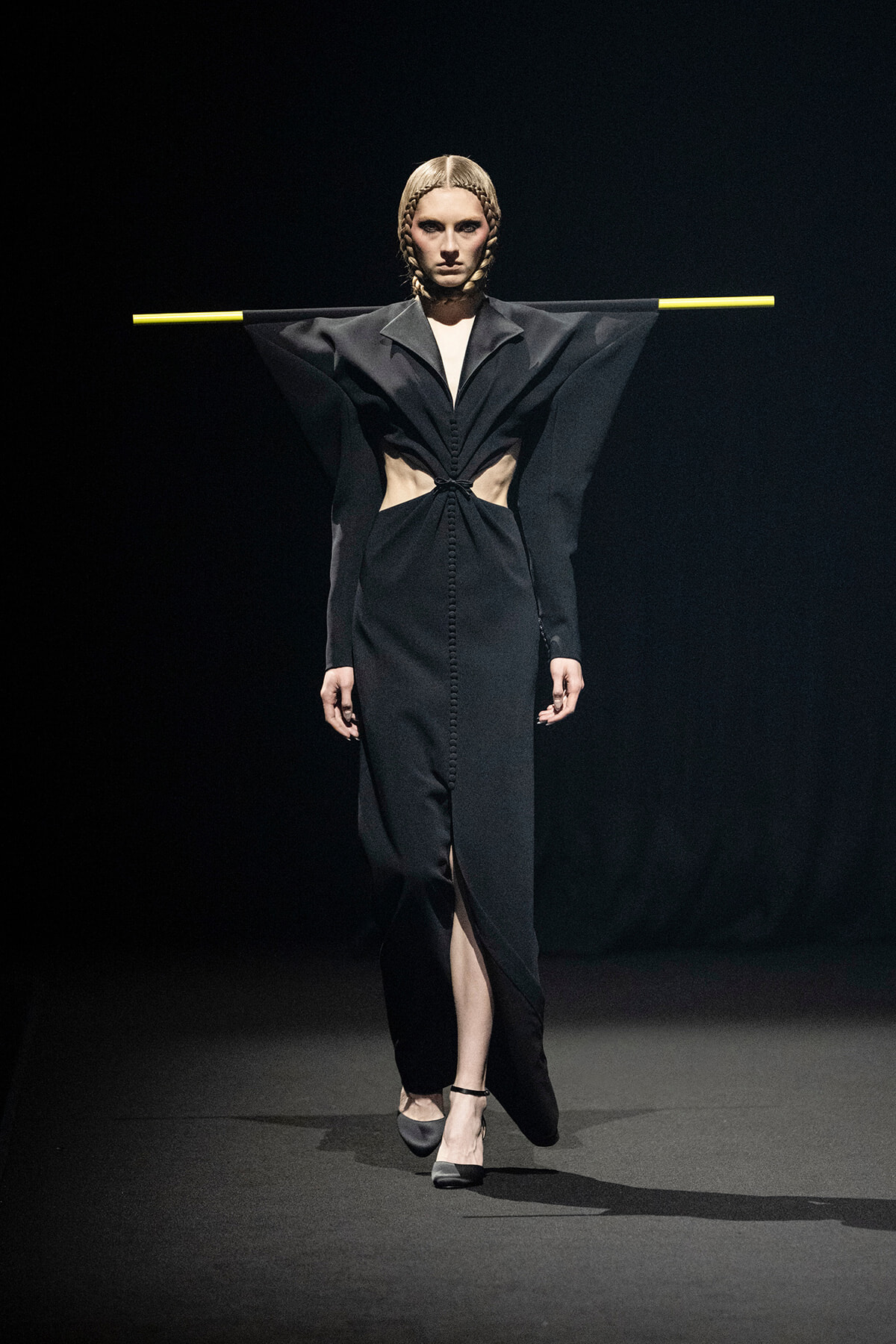 Model walking a fashion show in a black, avant-garde gown with wing-like sleeves, cutouts at the waist, and a front slit; hair styled in braids and arms stretched out with yellow bars across the sleeves.