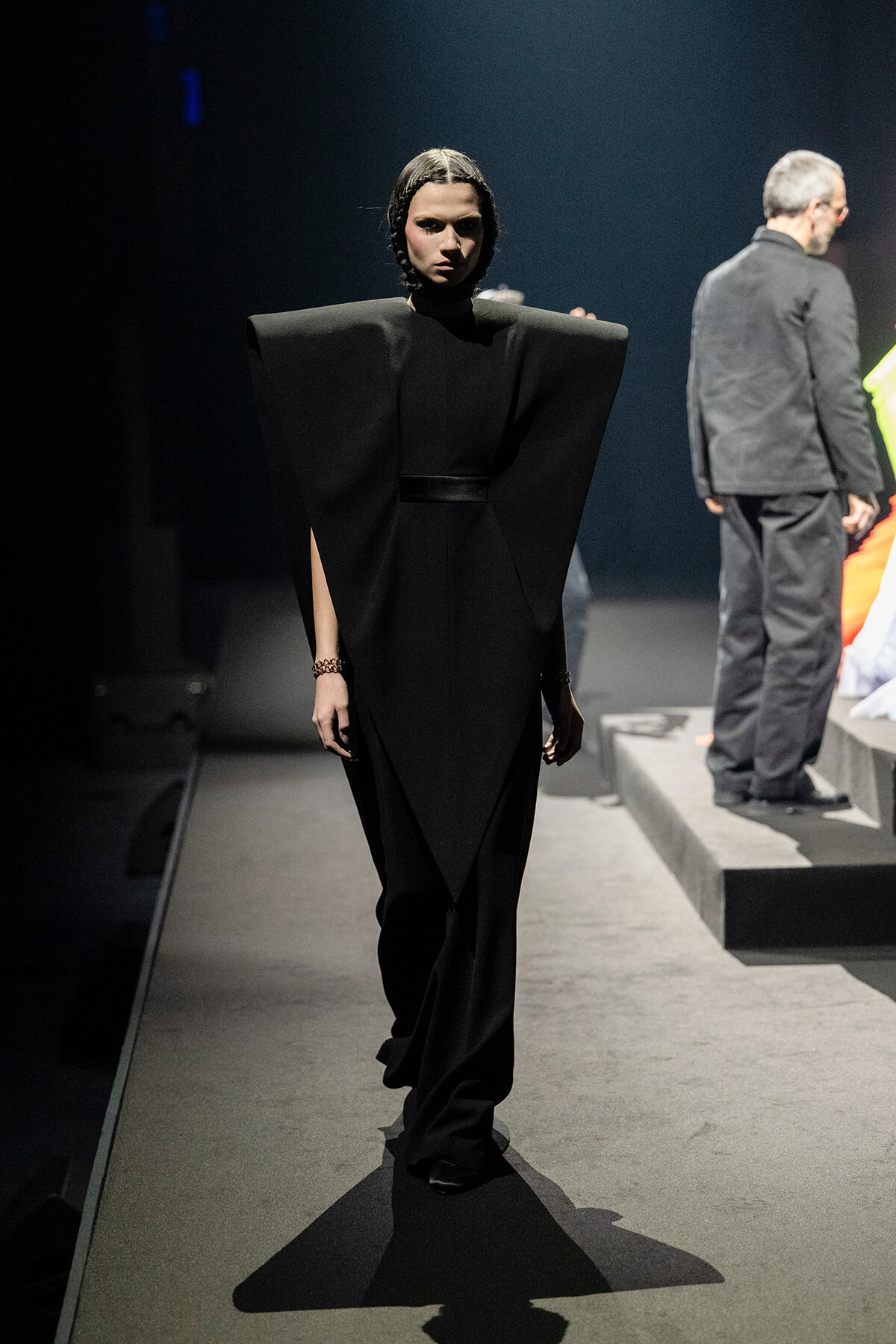 Model walks the runway in a black, sculptural gown with oversized, angular shoulders during a fashion show on a dimly lit catwalk.