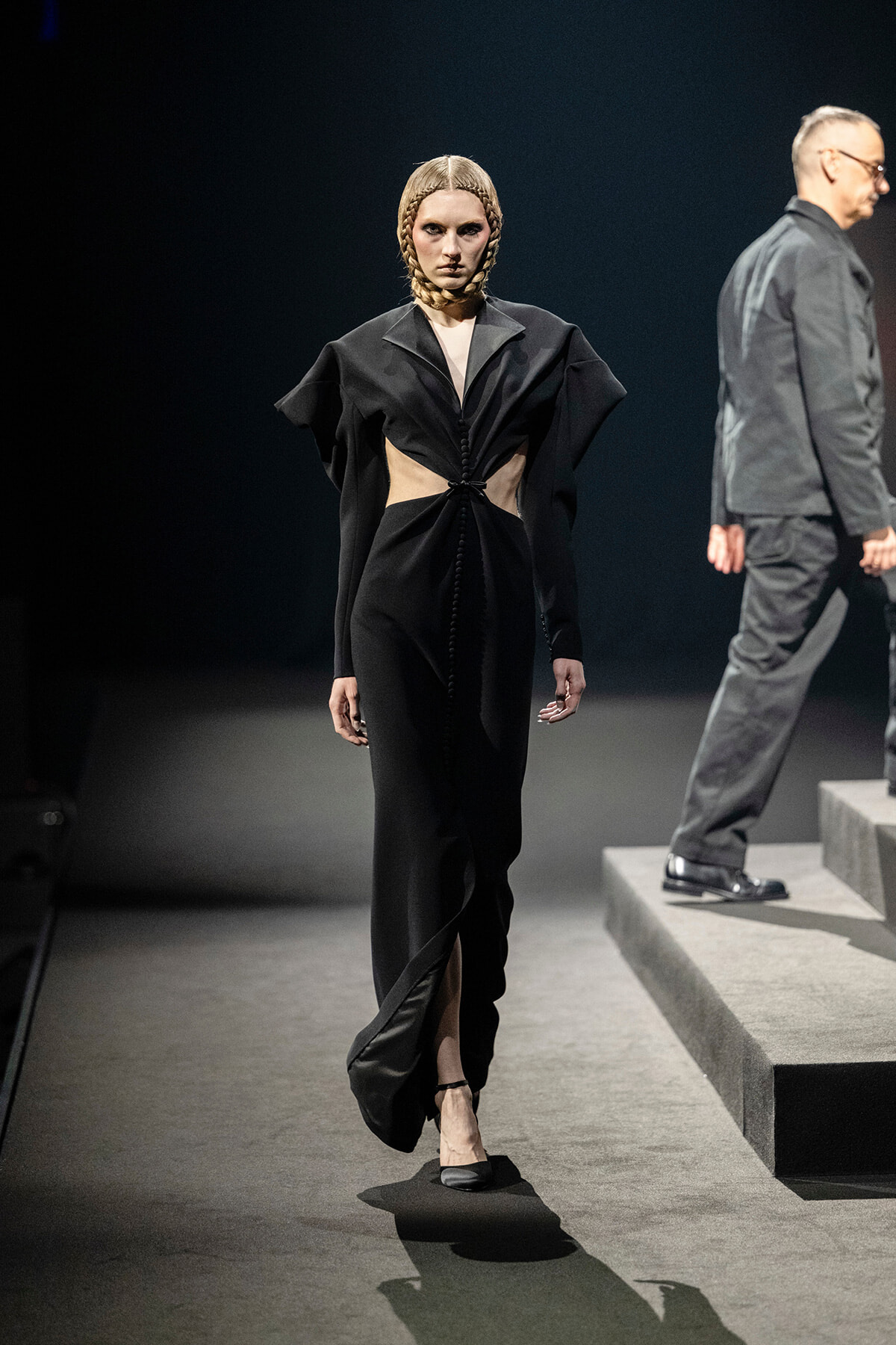 Model walking a runway in a sculptural black gown with waist cutouts and dramatic shoulders, braided hair, and black flats.