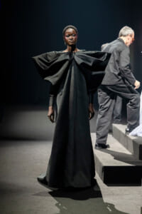 Model on a runway wearing a voluminous black gown with sculptural shoulders, standing center stage.