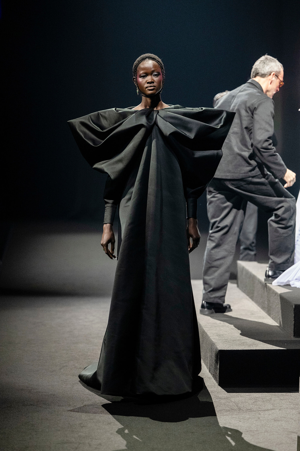 Model on a runway wearing a voluminous black gown with sculptural shoulders, standing center stage.