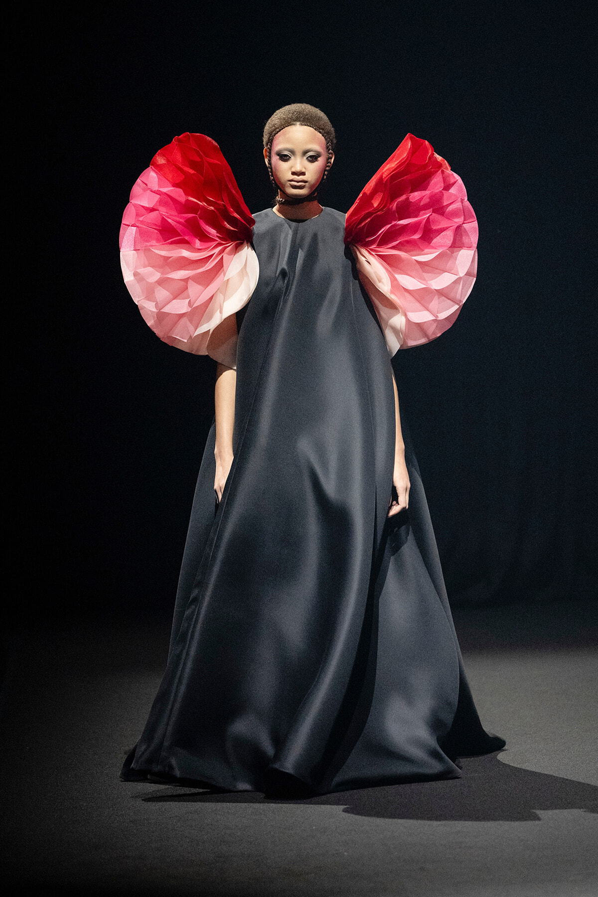 Model walks the runway wearing a dark, floor-length gown with oversized pink papercraft sleeves on each arm, set against a black stage backdrop.