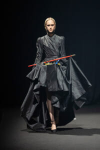 Model walking a dark runway in a dramatic black gown with a sculpted, voluminous skirt and braided hair, holding bright red and blue staff-like rods across the midsection.