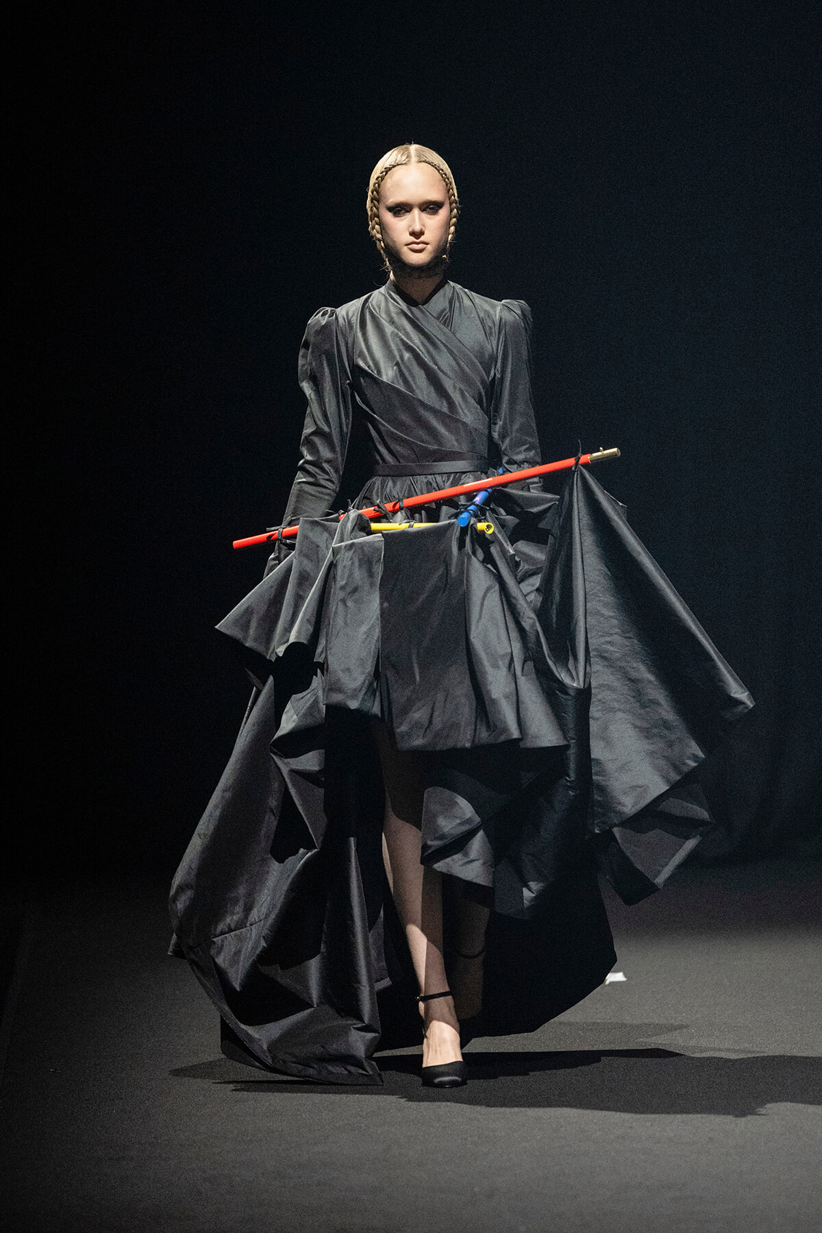 Model walking a dark runway in a dramatic black gown with a sculpted, voluminous skirt and braided hair, holding bright red and blue staff-like rods across the midsection.