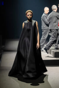 Model wearing a floor-length black satin cape-style gown walking the runway, with backstage crew blurred in the background.