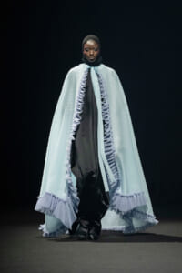 Model walks the runway in a pale blue cape with ruffled trim over a black outfit.