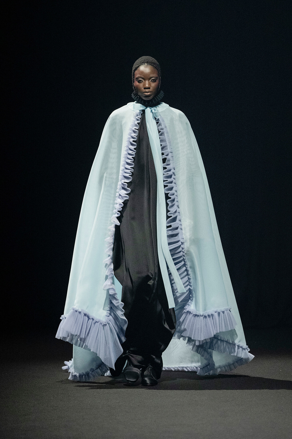 Model walks the runway in a pale blue cape with ruffled trim over a black outfit.