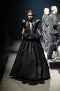 Female model walking a dark runway in a voluminous black gown with structured shoulders and deep V neckline.