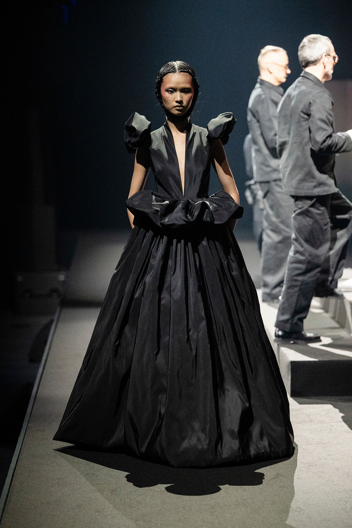 Female model walking a dark runway in a voluminous black gown with structured shoulders and deep V neckline.