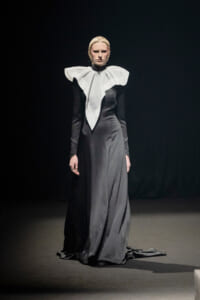 Model on a runway wearing a long black gown with a dramatic white oversized ruffled collar.