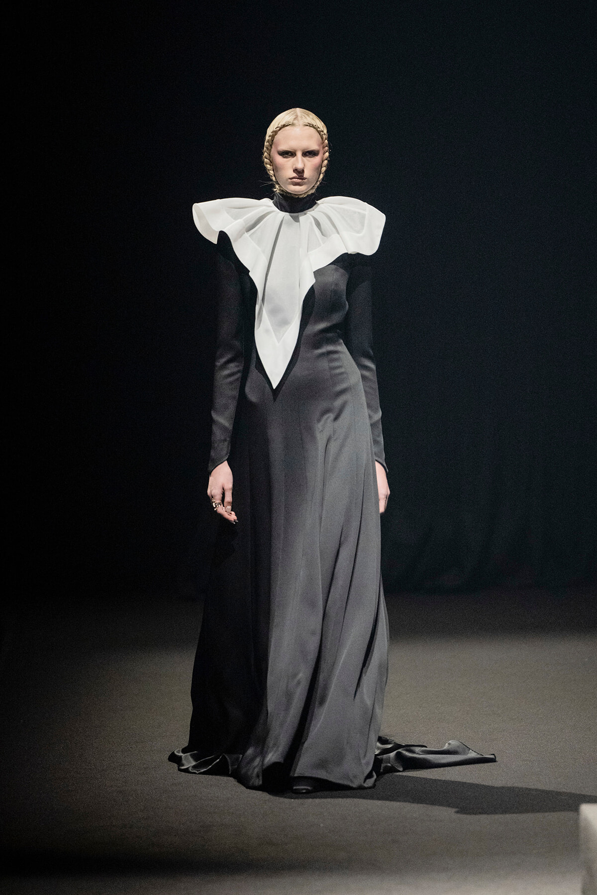 Model on a runway wearing a long black gown with a dramatic white oversized ruffled collar.