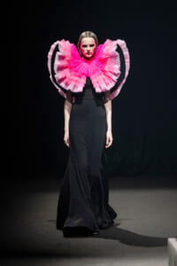 Model wearing a dramatic hot-pink ruff collar over a long black gown on a runway, dark background behind.