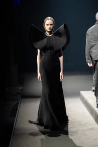 Female model on a runway wearing an all-black gown with oversized sculptural shoulder pieces.
