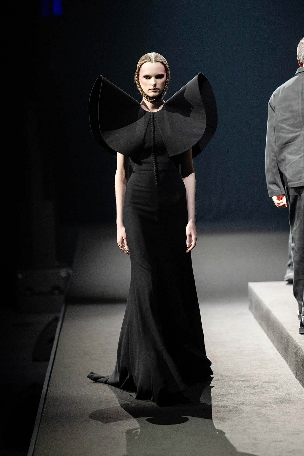 Female model on a runway wearing an all-black gown with oversized sculptural shoulder pieces.