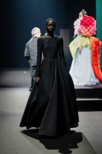 Black woman in a floor-length textured black gown with a full skirt on a fashion runways; colorful outfits and a person in the background on stage shift the focus to the model.