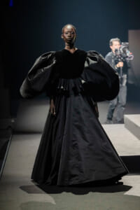 Runway model in a dramatic black gown with oversized puff sleeves and a full skirt, walking the catwalk as a cameraman films in the background.