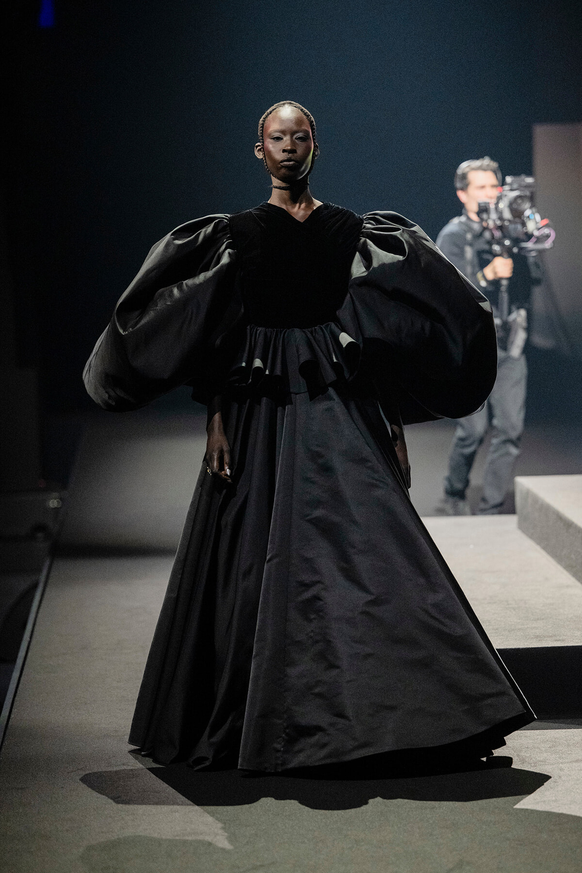 Runway model in a dramatic black gown with oversized puff sleeves and a full skirt, walking the catwalk as a cameraman films in the background.