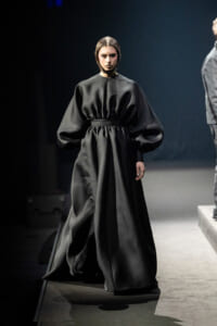 Model walking a fashion runway in a dramatic, voluminous black gown with a cinched waist and exaggerated sleeves.