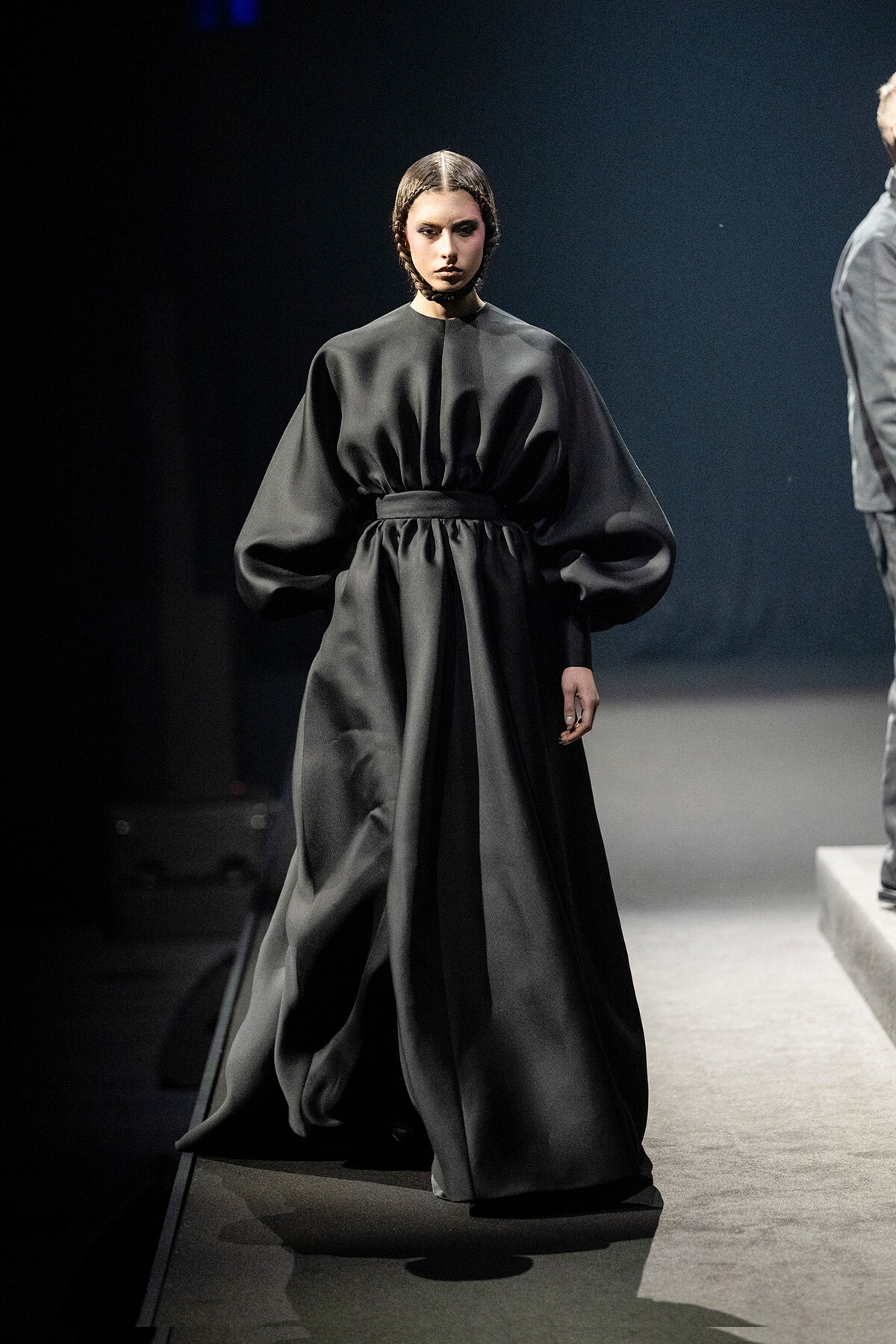 Model walking a fashion runway in a dramatic, voluminous black gown with a cinched waist and exaggerated sleeves.