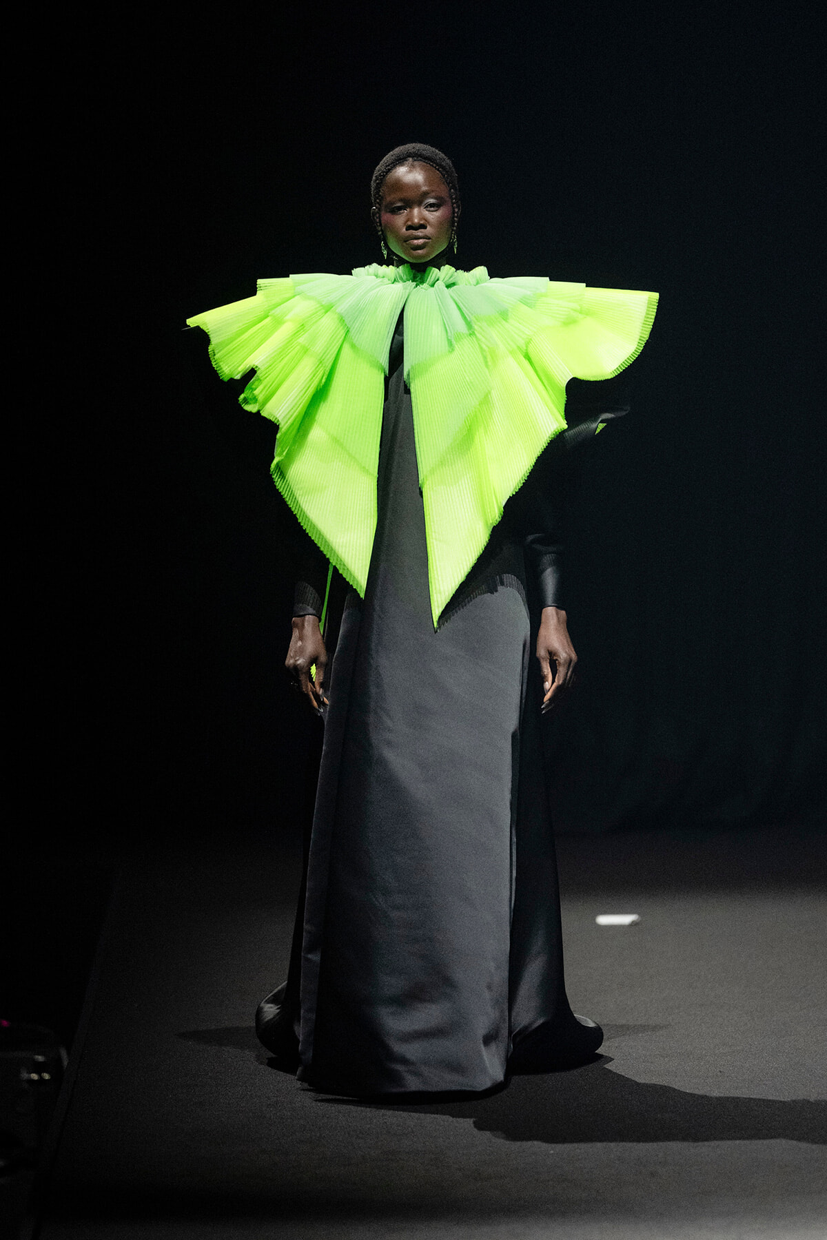 Model on runway wearing a neon lime-green sculptural cape over a dark gown against a black backdrop.