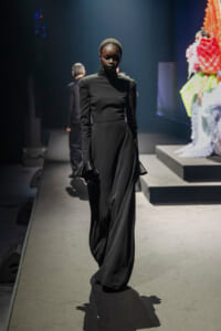 Model walks the runway in a sleek black high-neck gown with dramatic flared cuffs.