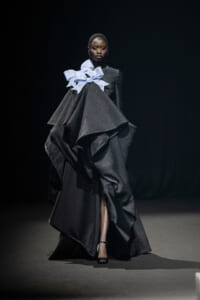 Model on a runway wearing a voluminous black textured gown with layered panels and a large pale blue bow at the neckline/shoulder area?