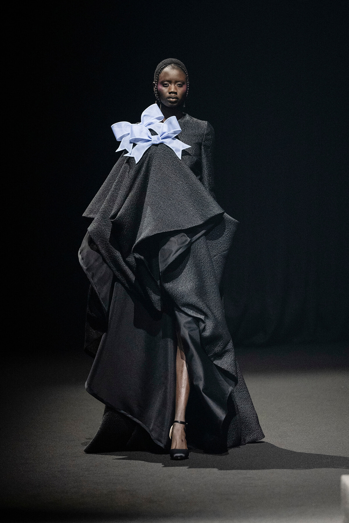 Model on a runway wearing a voluminous black textured gown with layered panels and a large pale blue bow at the neckline/shoulder area?