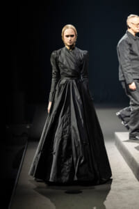 Model on a runway wearing a long black satin Victorian-style gown with a structured bodice and braided hair.