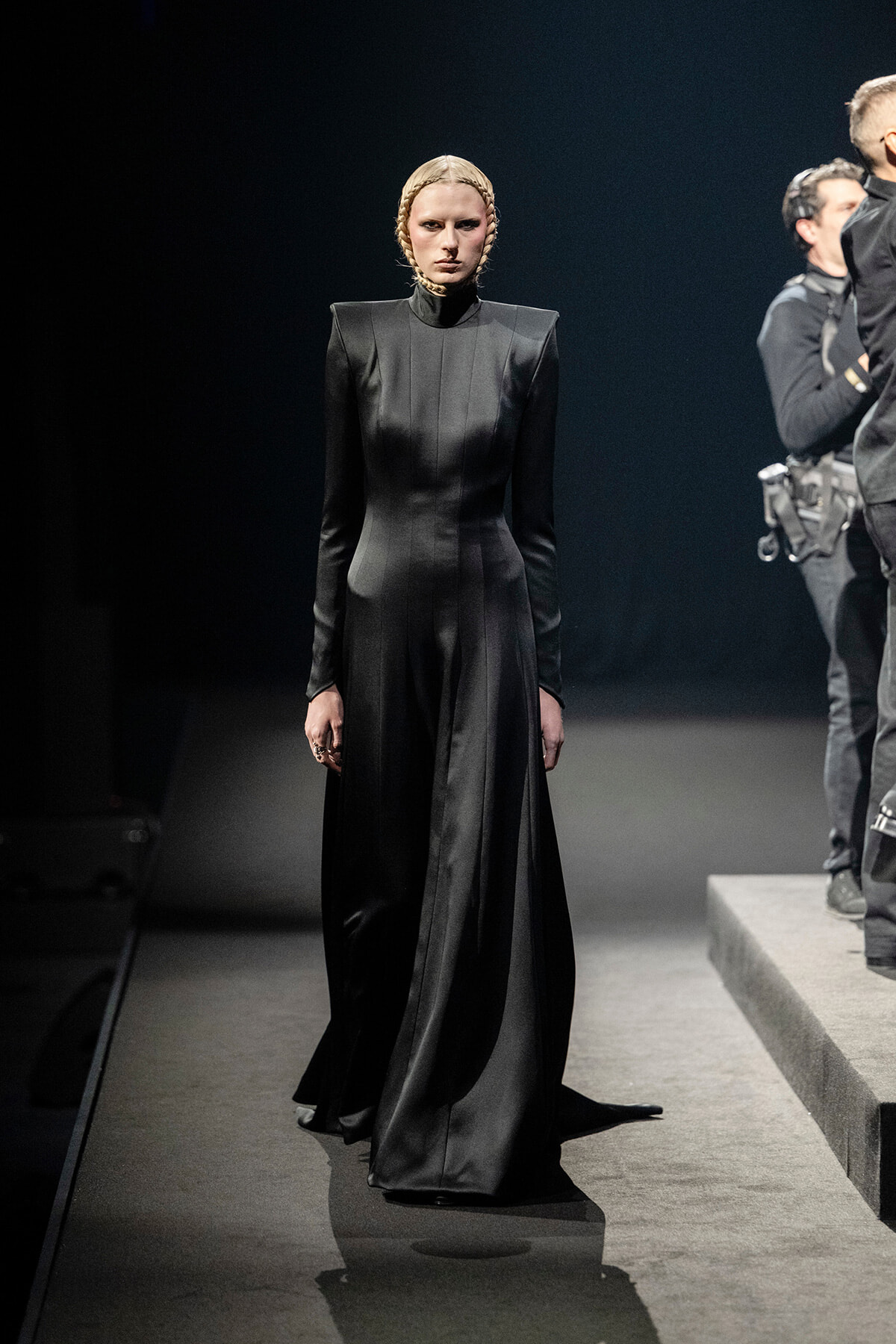 Model walks the runway in a floor-length black high-neck gown with structured shoulders and braided blonde hair, spotlight on stage.