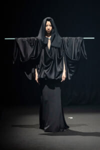 Model in a dramatic black, draped gown with wide sleeves, arms extended on a horizontal bar against a dark stage backdrop.