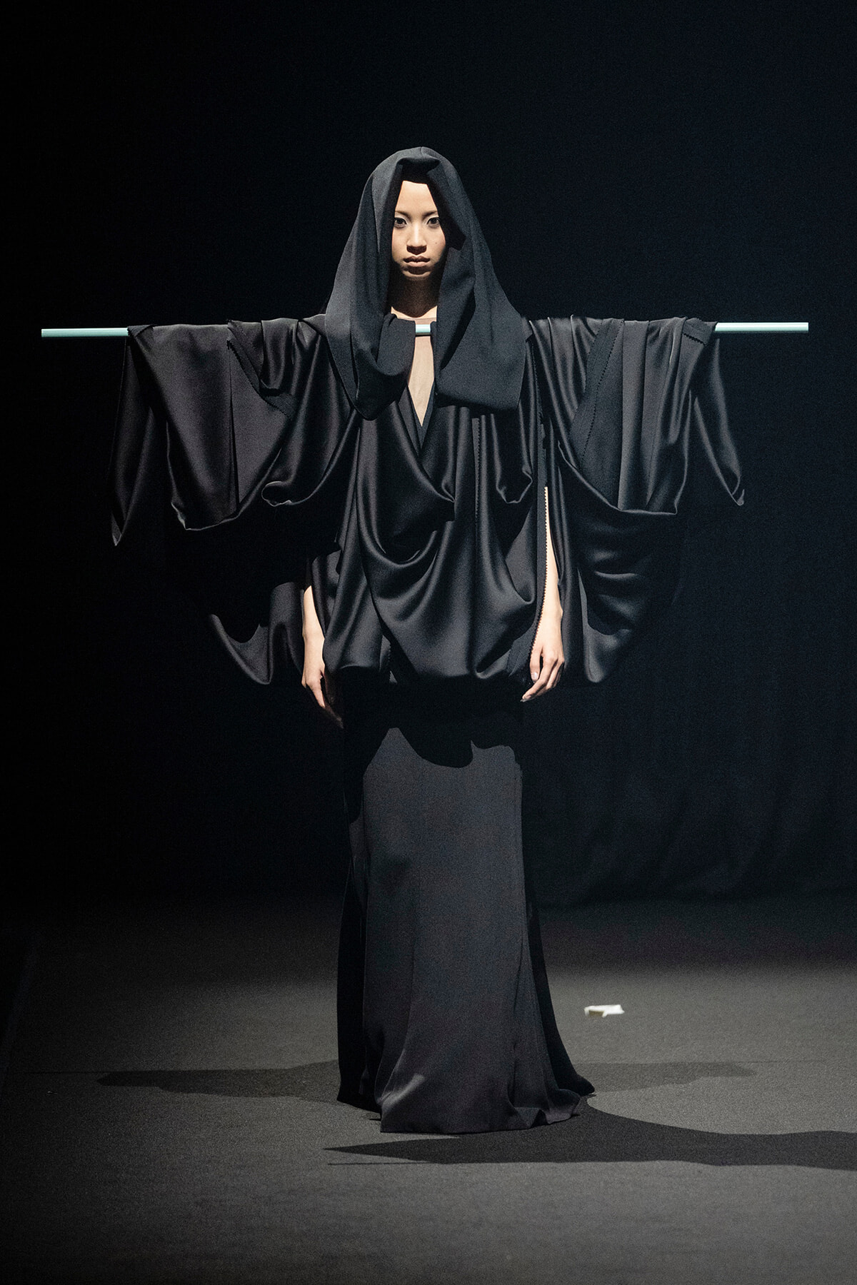 Model in a dramatic black, draped gown with wide sleeves, arms extended on a horizontal bar against a dark stage backdrop.