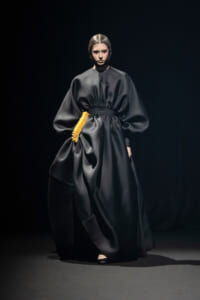 Full-length black satin gown with oversized sleeves and a bright orange waist accessory on a studio runway, model posing.