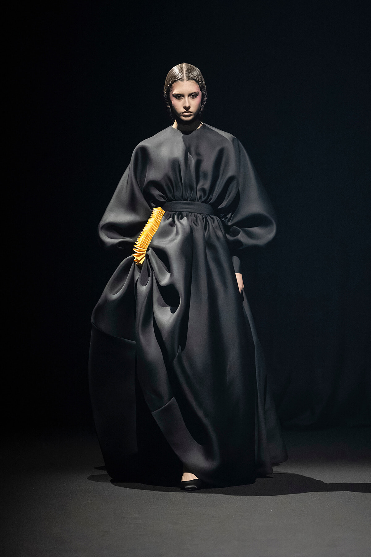 Full-length black satin gown with oversized sleeves and a bright orange waist accessory on a studio runway, model posing.