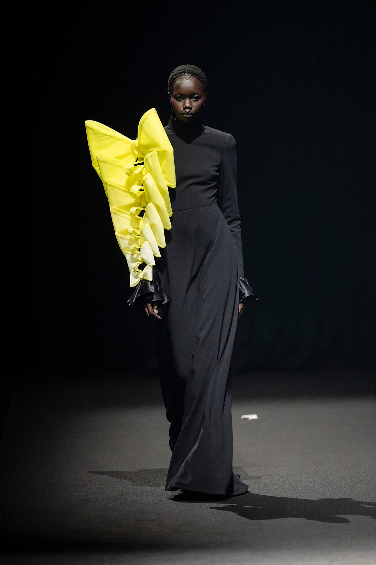 Model on a runway wearing a long black dress with a large neon yellow sculptural ruffle along the left arm/shoulder, dramatic contrast and pose.