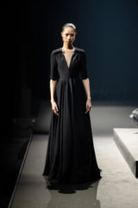 Model on a runway wearing a floor-length black gown with a sheer V-neck panel and three-quarter sleeves, under spotlight.