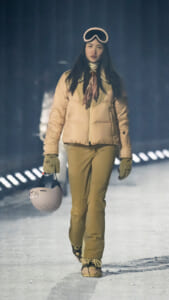 Woman walking on a snowy runway in a beige puffer jacket, ski goggles on her head, scarf, gloves, and holding a pale pink ski helmet