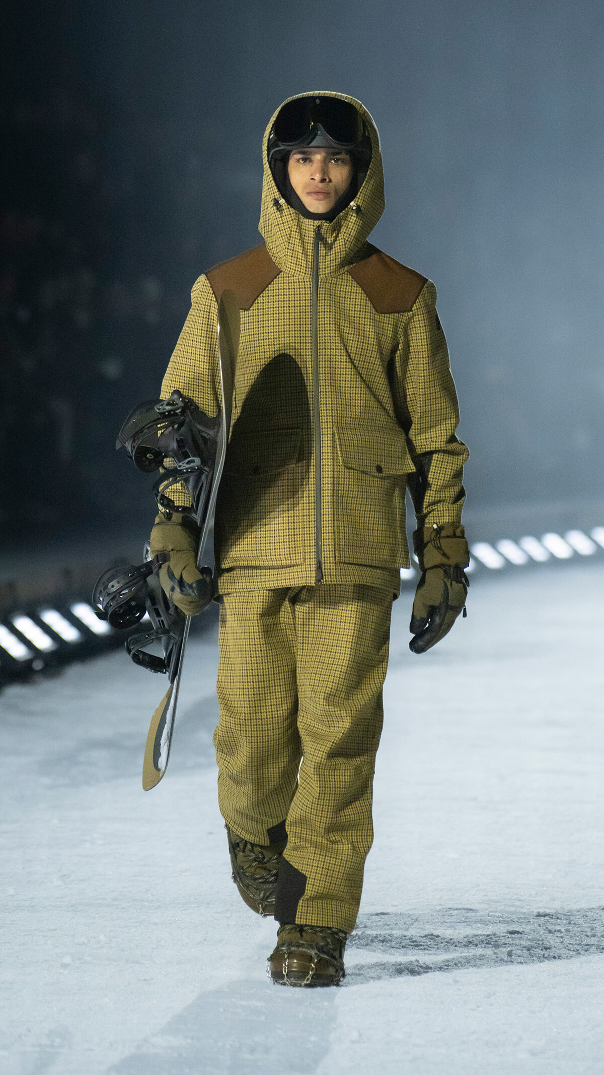 Model walking on a snowy runway in a yellow plaid snowsuit with hood and goggles, carrying a snowboard under one arm.