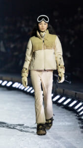 Woman models a beige fringe-vested winter jacket on a snow runway, wearing ski goggles and holding ski poles.