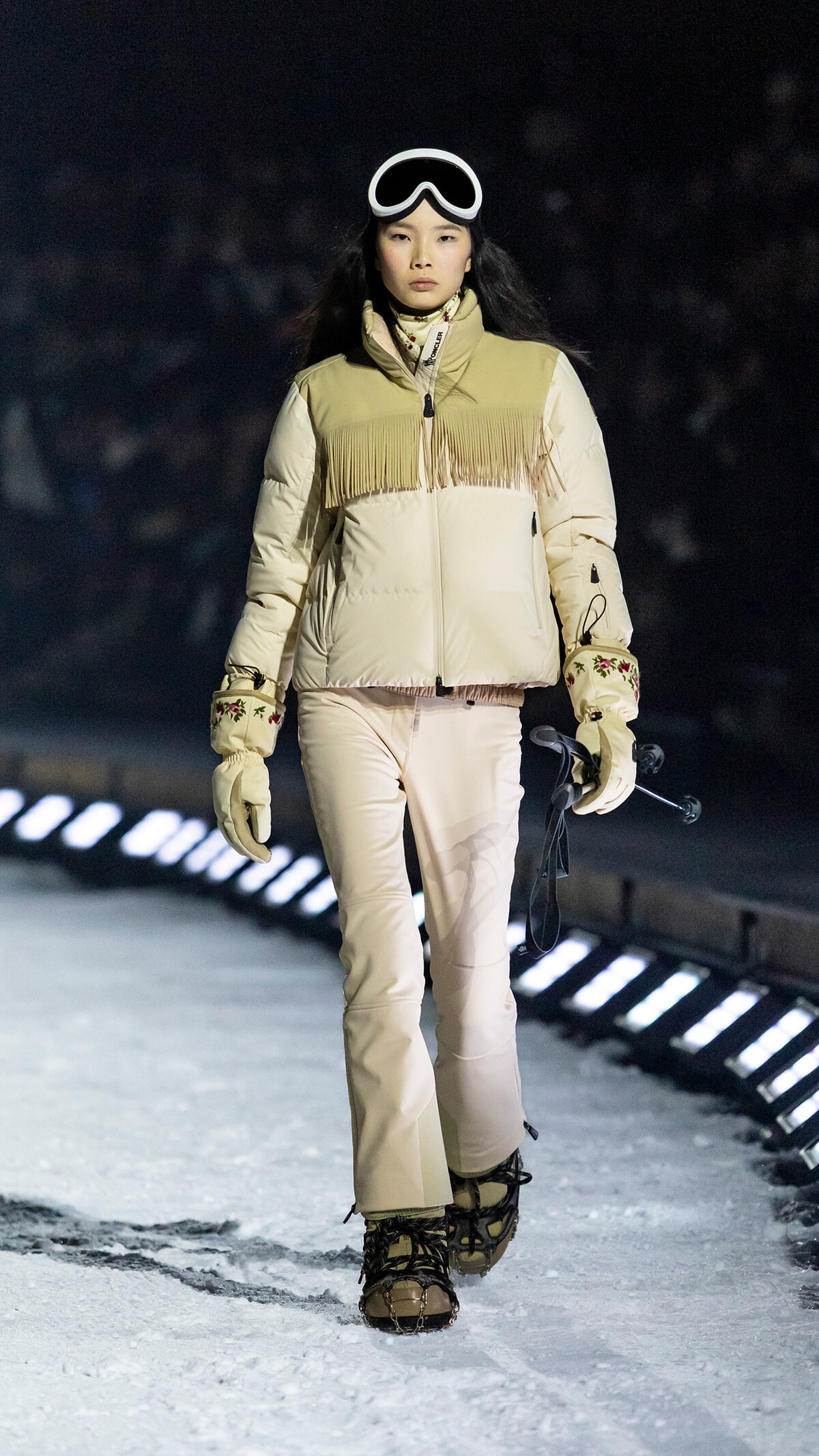 Woman models a beige fringe-vested winter jacket on a snow runway, wearing ski goggles and holding ski poles.