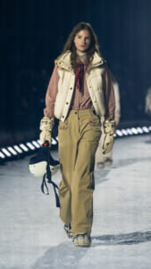 Model walking on a snowy runway in a beige embroidered outfit with a white puffer vest, plaid shirt, gloves, and snow boots, holding a white helmet.