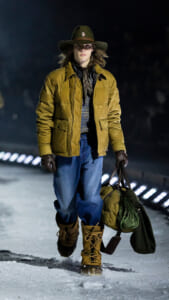 Person walking in snow wearing a tan padded jacket, jeans, brown boots, and a wide-brim hat, carrying a duffel bag and climbing gear; night scene.”,