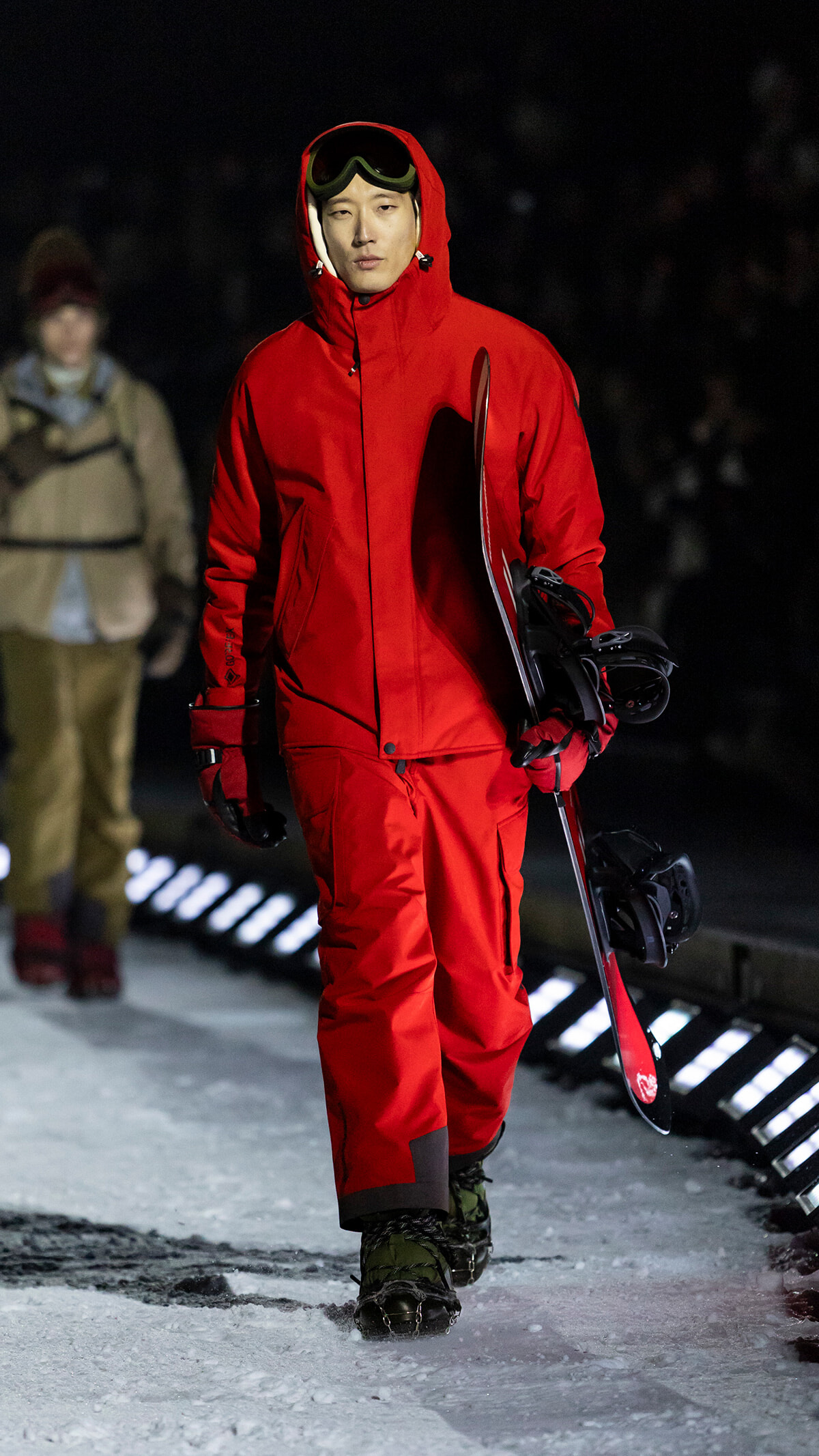 Person wearing a bright red snowsuit and ski goggles walks on a snowy runway, carrying a snowboard under their arm.