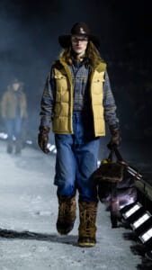 Model walking on an icy runway at night in a tan puffer vest, plaid shirt, blue jeans, fur-lined boots, and a wide-brim hat, carrying a duffel bag.